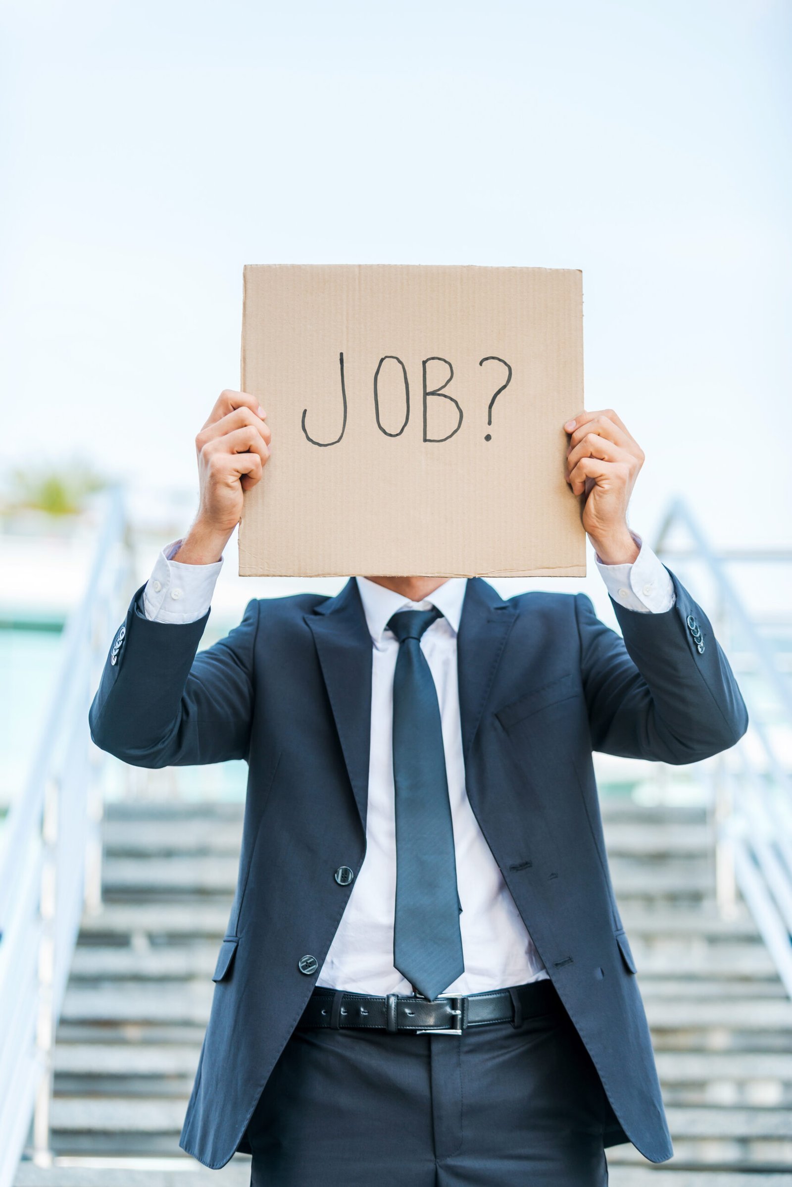 i need a job! man in formalwear holding poster with job text message in front of his face while standing outdoors and against staircase